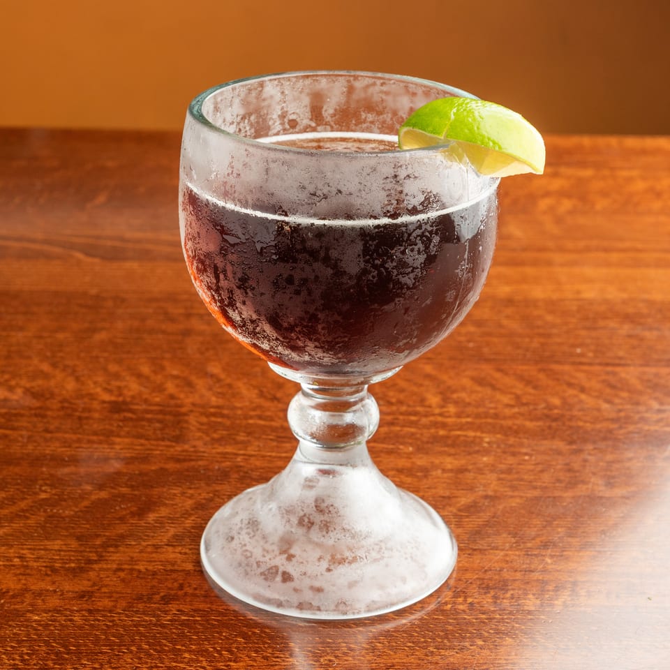 A frosty glass of dark soda with a lime wedge sits on a wooden table.
