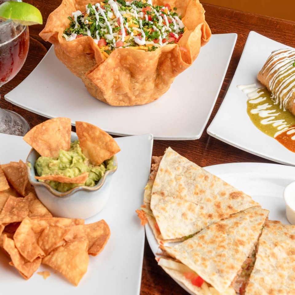 A table laden with Mexican dishes: a taco salad, a burrito, guacamole with chips, and quesadillas.