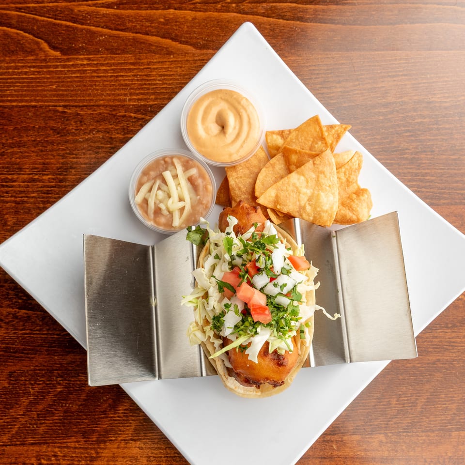 Overhead view of a fish taco with shredded cabbage, diced tomatoes, and cilantro, served with chips, beans, and sauce.