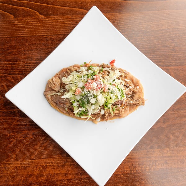 A Mexican sopes dish with shredded pork, cabbage, tomatoes, and cheese on a white plate.