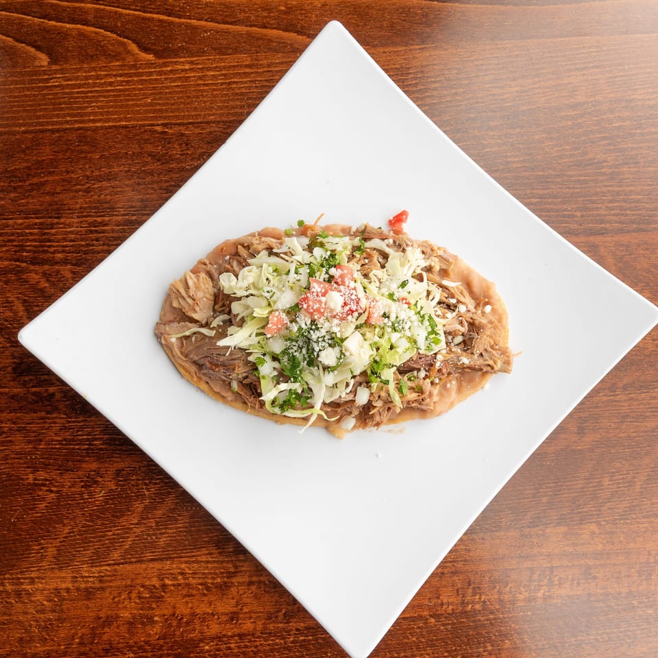 A Mexican sopes dish with shredded pork, cabbage, tomatoes, and cheese on a white plate.