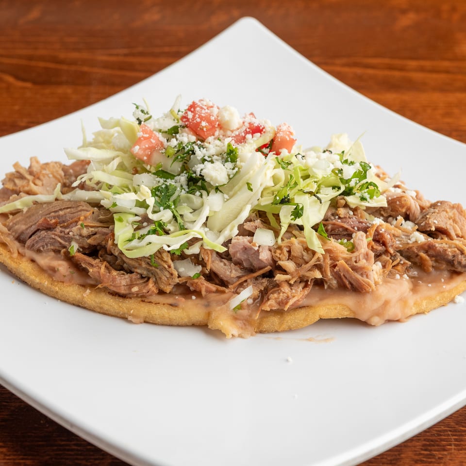 A Mexican sopes with shredded pork, refried beans, cabbage, tomatoes, and crumbled cheese.