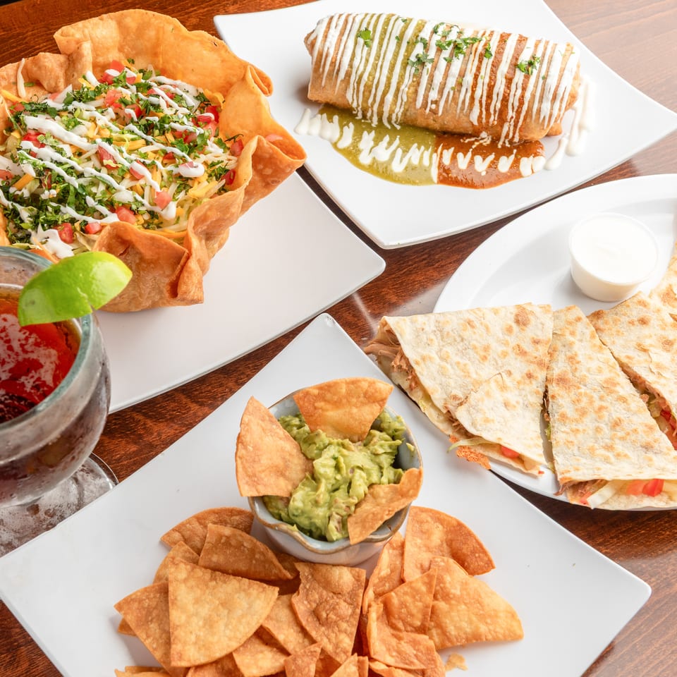 A taco salad, burrito, quesadillas, guacamole with chips, and a drink with lime on a wooden table.