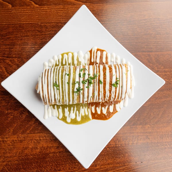 A burrito covered in white, green, and red sauces, garnished with cilantro, on a white plate.