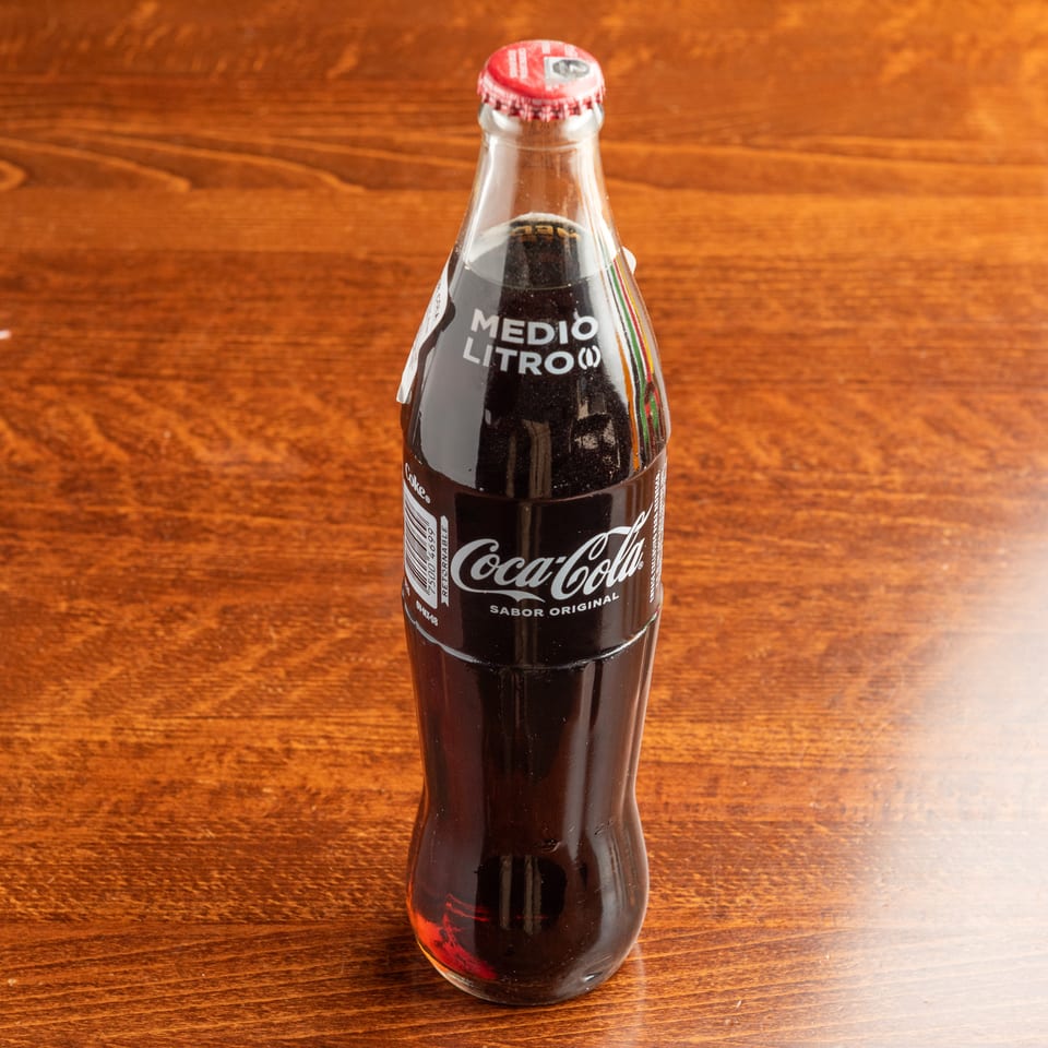 A glass bottle of Mexican Coca-Cola, labeled "Medio Litro" and "Sabor Original," on a wooden table.