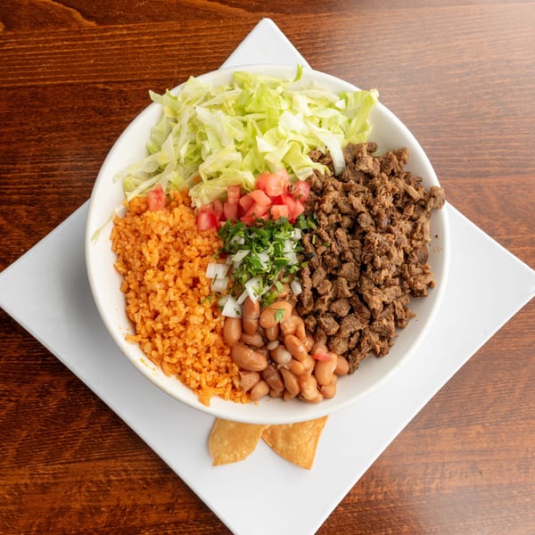 A white bowl filled with rice, beans, diced beef, lettuce, tomatoes, and onions on a wooden table.