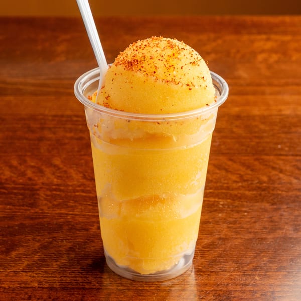 A mango chamoyada, a Mexican frozen drink, in a clear plastic cup with a white spoon.