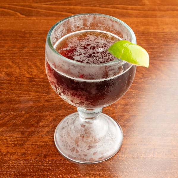 A frosty glass of dark red beverage with a lime wedge on a wooden table.