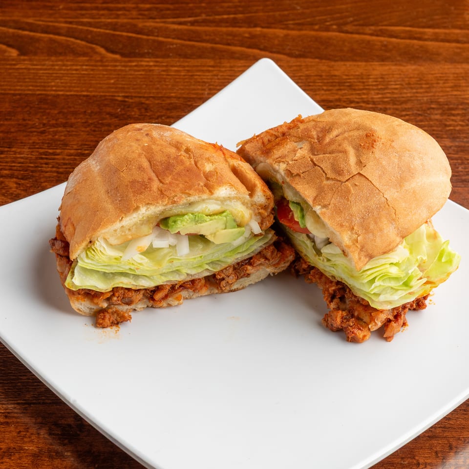 A torta sandwich cut in half, revealing layers of meat, lettuce, avocado, and onions on a white plate.